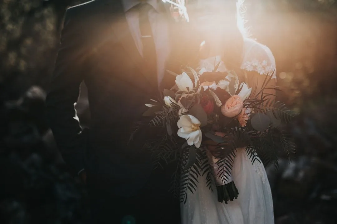 A wedding couple stands in golden sunlight holding a bouquet of white flowers and ferns at sunset.