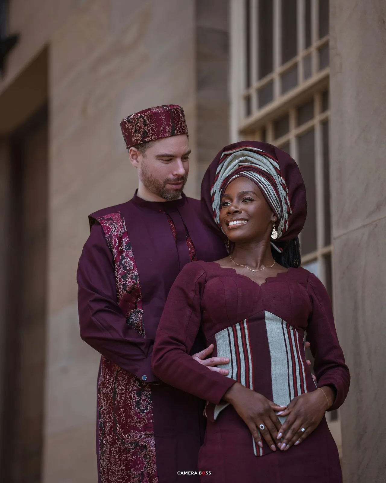 A couple in coordinating burgundy traditional attire pose together, with her wearing a gele headwrap and him in ethnic formal wear.