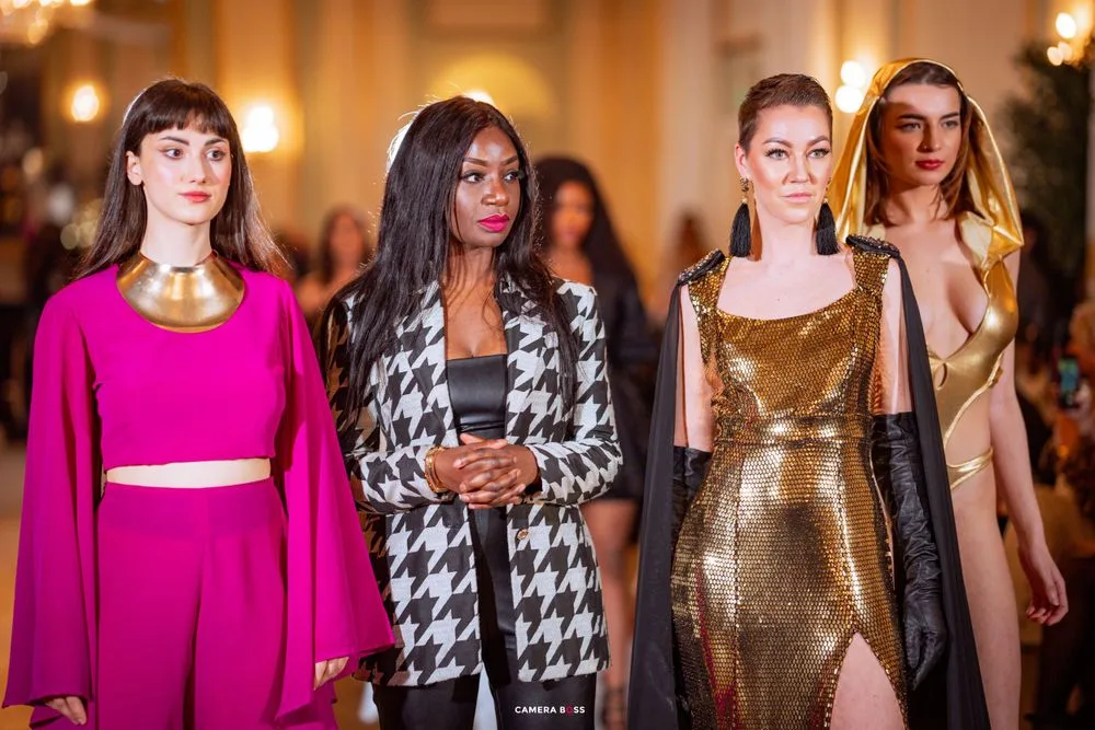 Fashion show models walk the runway wearing bold designs including a pink crop top, houndstooth jacket, and metallic gold dress.