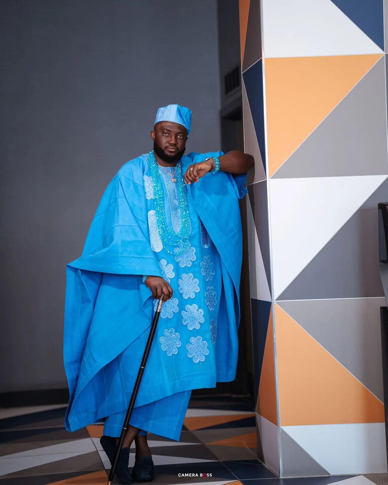 Traditional African male attire featuring a bright blue agbada with embroidered details and matching cap against geometric wall art.