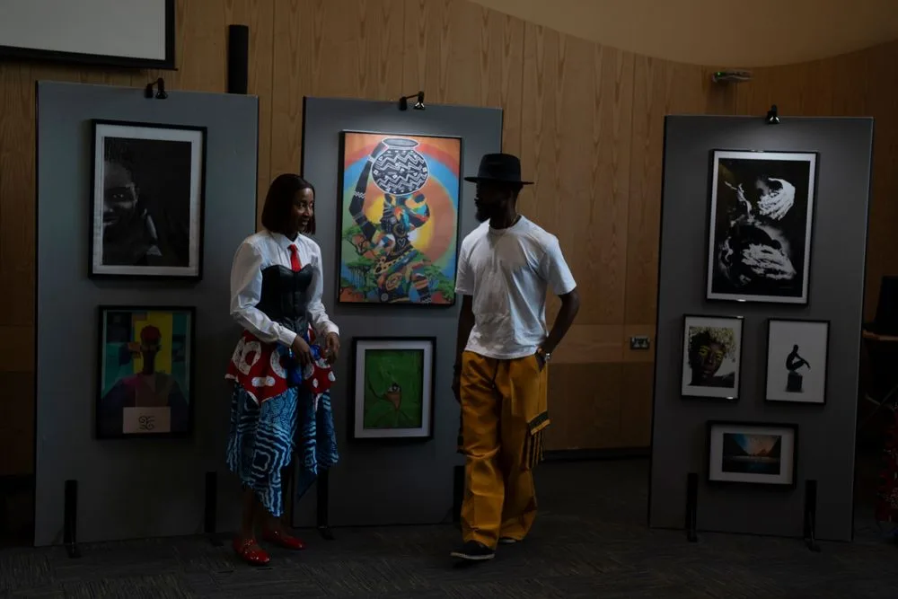 Two people converse at an art gallery exhibition featuring colorful paintings and black-and-white photographs on display panels.