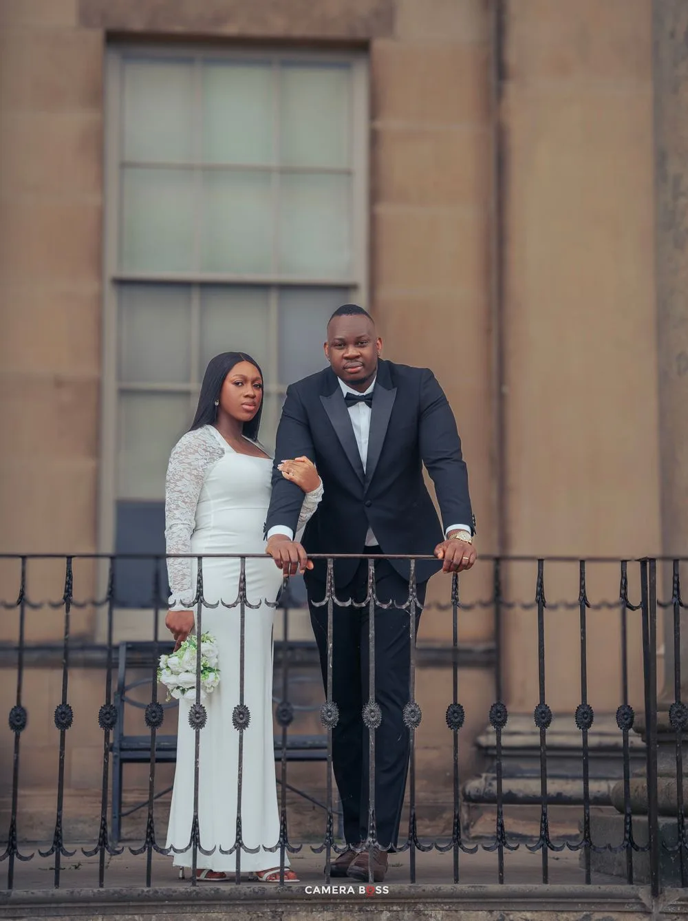 A couple in formal wedding attire stands on a balcony with iron railings against a stone building backdrop.