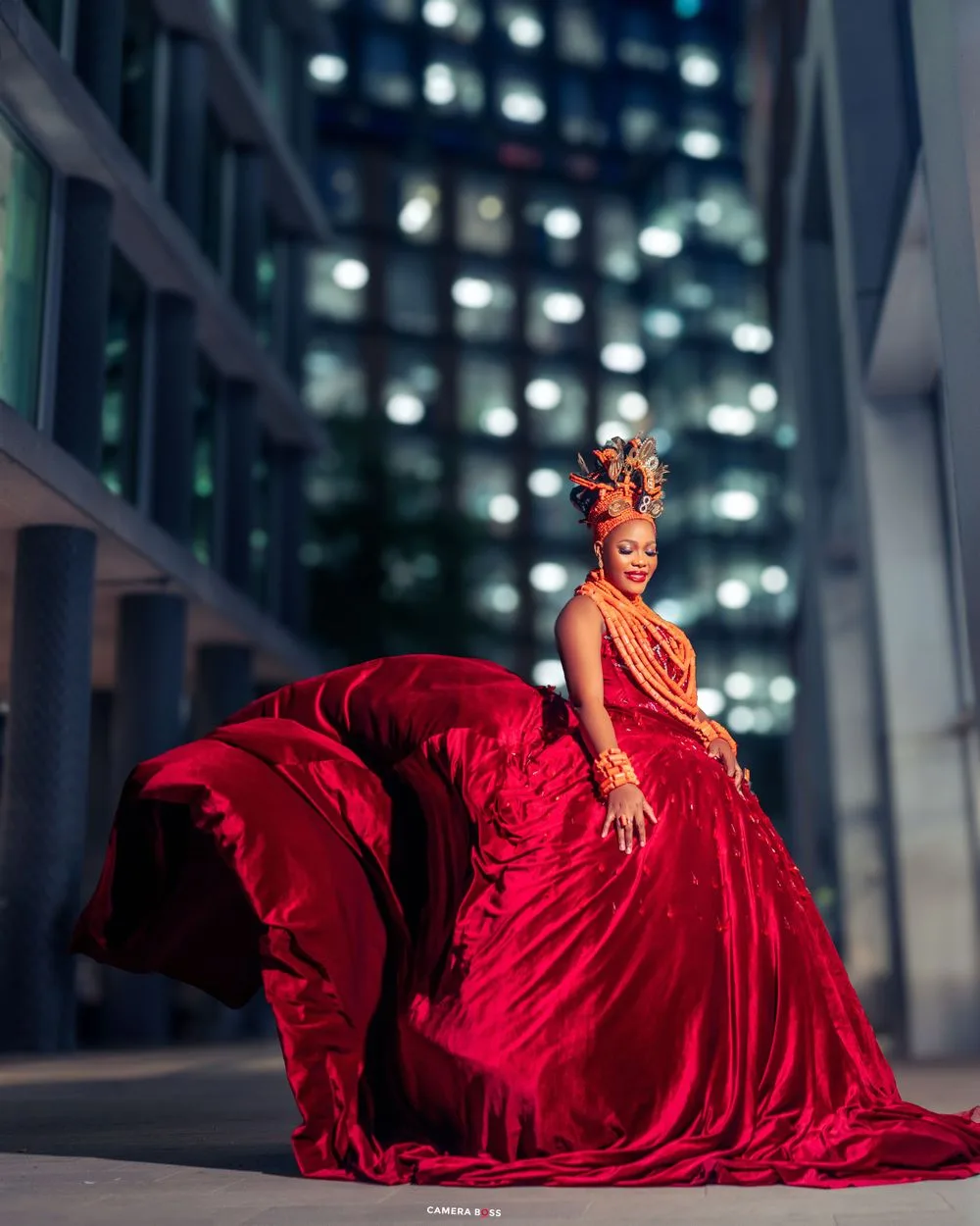 A model in a flowing red ballgown poses dramatically against a modern building with glowing windows at night.