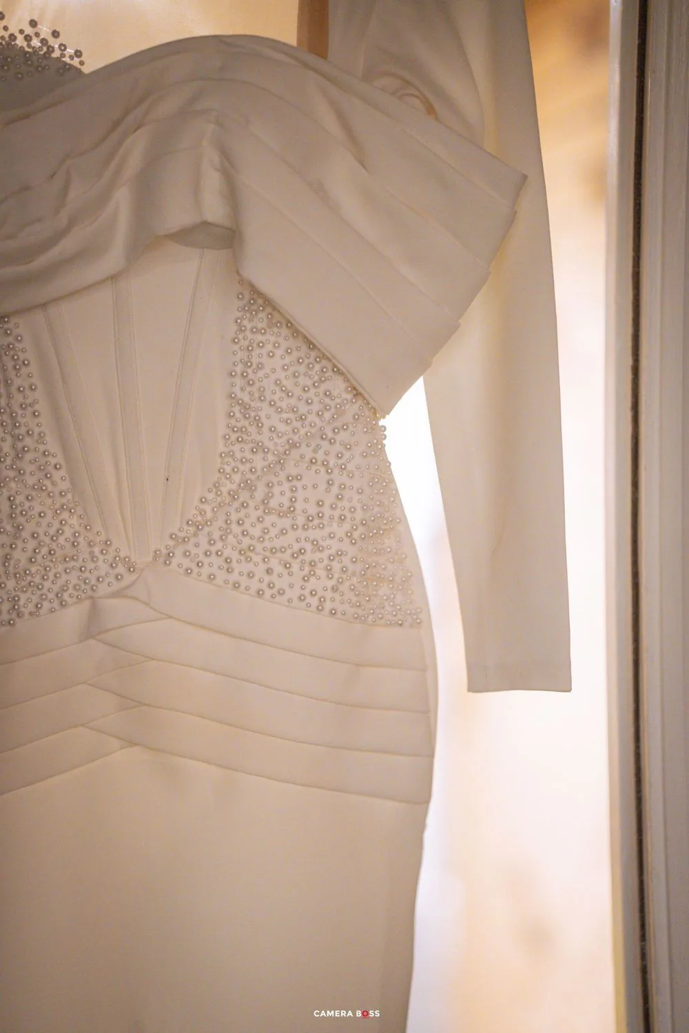 A detailed shot of an elegant white wedding dress with beaded bodice and ruched details hanging against a softly lit wall.