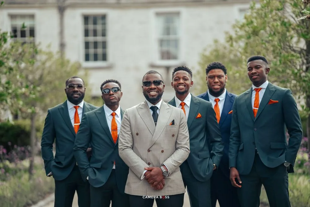 A wedding party wearing navy suits with orange ties poses together outdoors with one member in a beige suit.