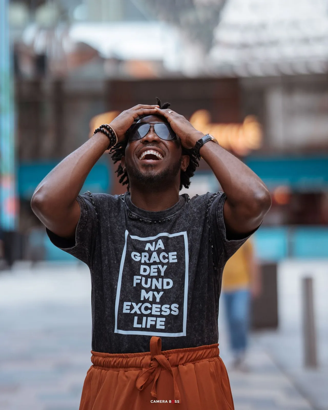 Person wearing a black graphic t-shirt and orange pants laughs joyfully on a city street.