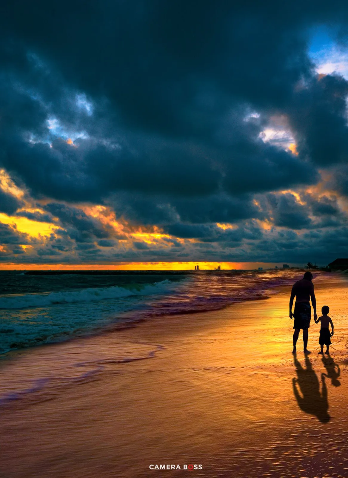 Two silhouettes walk along a beach at sunset with dramatic stormy clouds and golden light reflecting on the wet sand.