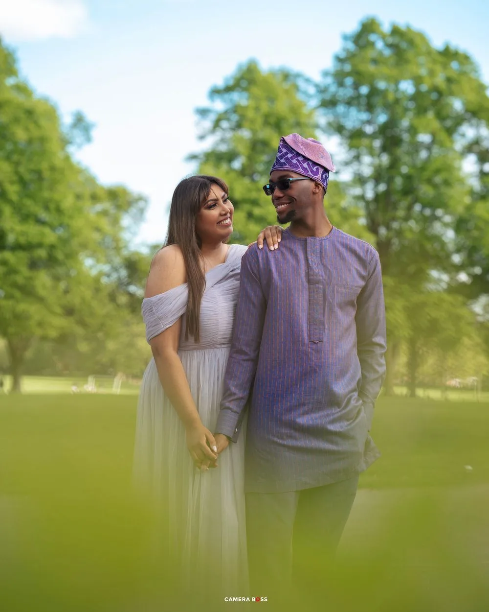 A couple in coordinated purple and white outfits share an intimate moment together in a sunlit park setting.
