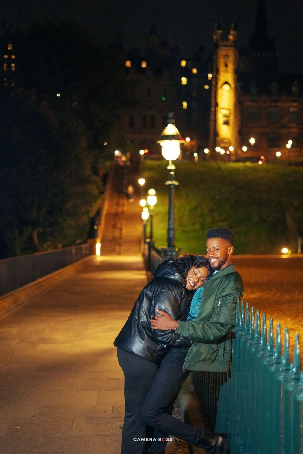 Two friends share a joyful moment on a lamp-lit pathway at night with historic buildings and towers in the background.
