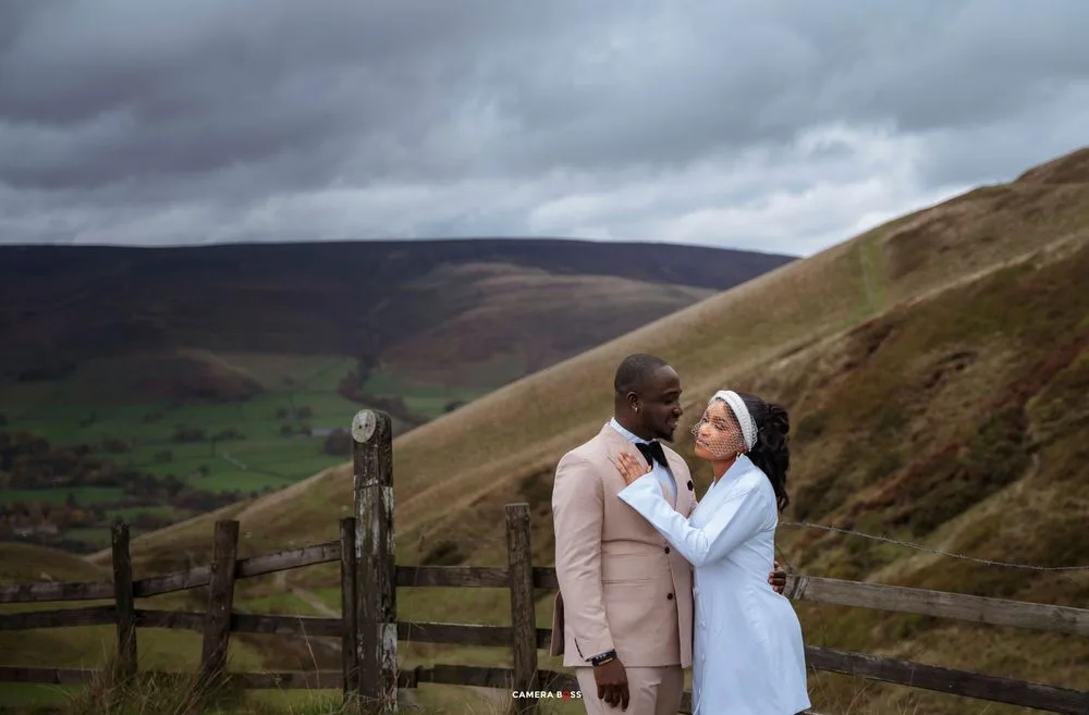 Couple sharing a romantic moment by a wooden fence with rolling green hills and dramatic cloudy skies.