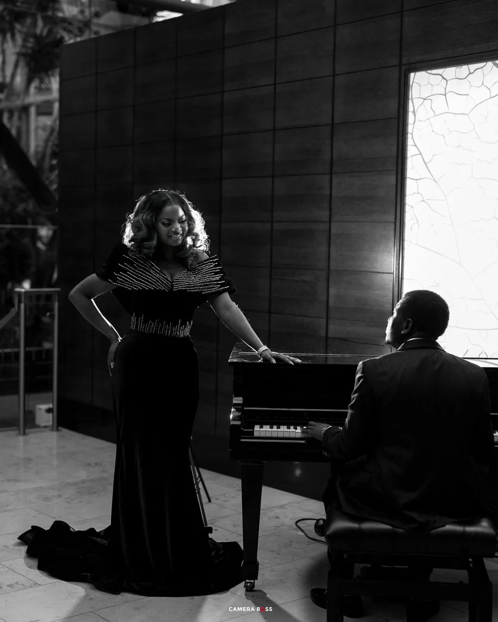Black and white photo of a woman in an elegant gown standing by a piano while a man plays in a formal setting.