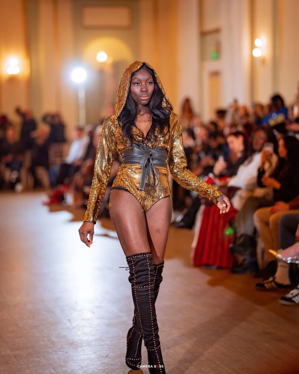 Fashion model walks runway in gold metallic hooded bodysuit and thigh-high boots at luxury fashion show.