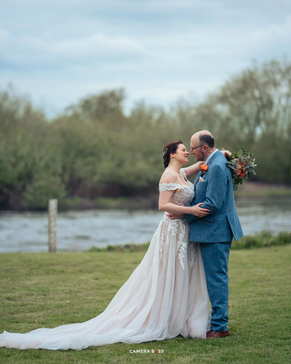 A couple shares a romantic moment by a lake on their wedding day, with the bride in a flowing off-shoulder gown.