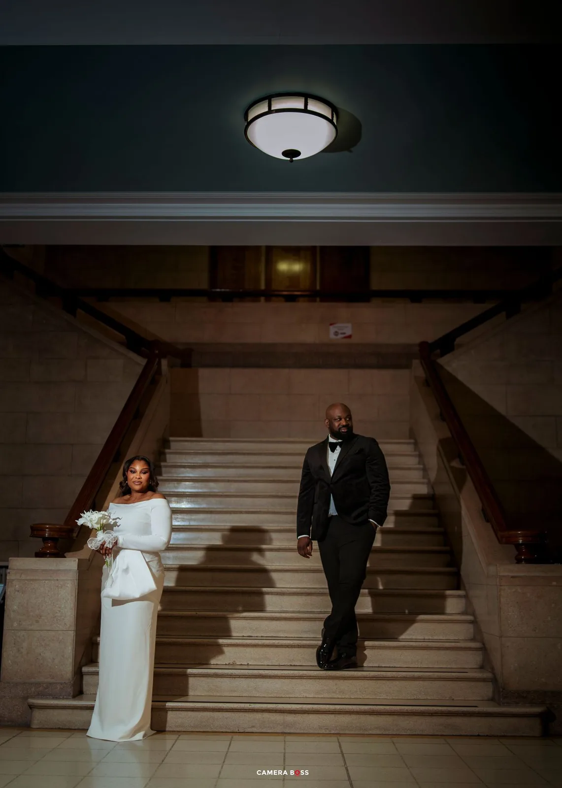 Bride and groom descend grand marble staircase, bride in sleek white gown with bouquet, groom in classic black tuxedo.