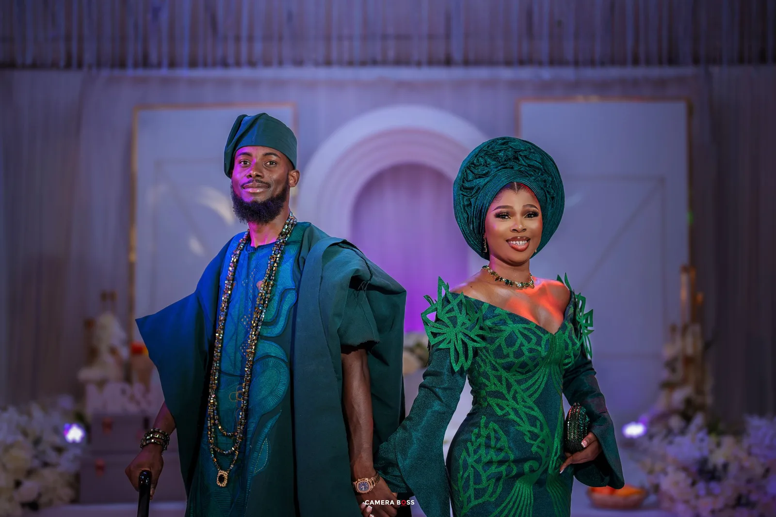 Two people wearing traditional Nigerian attire pose during a formal event with emerald green and blue outfits.