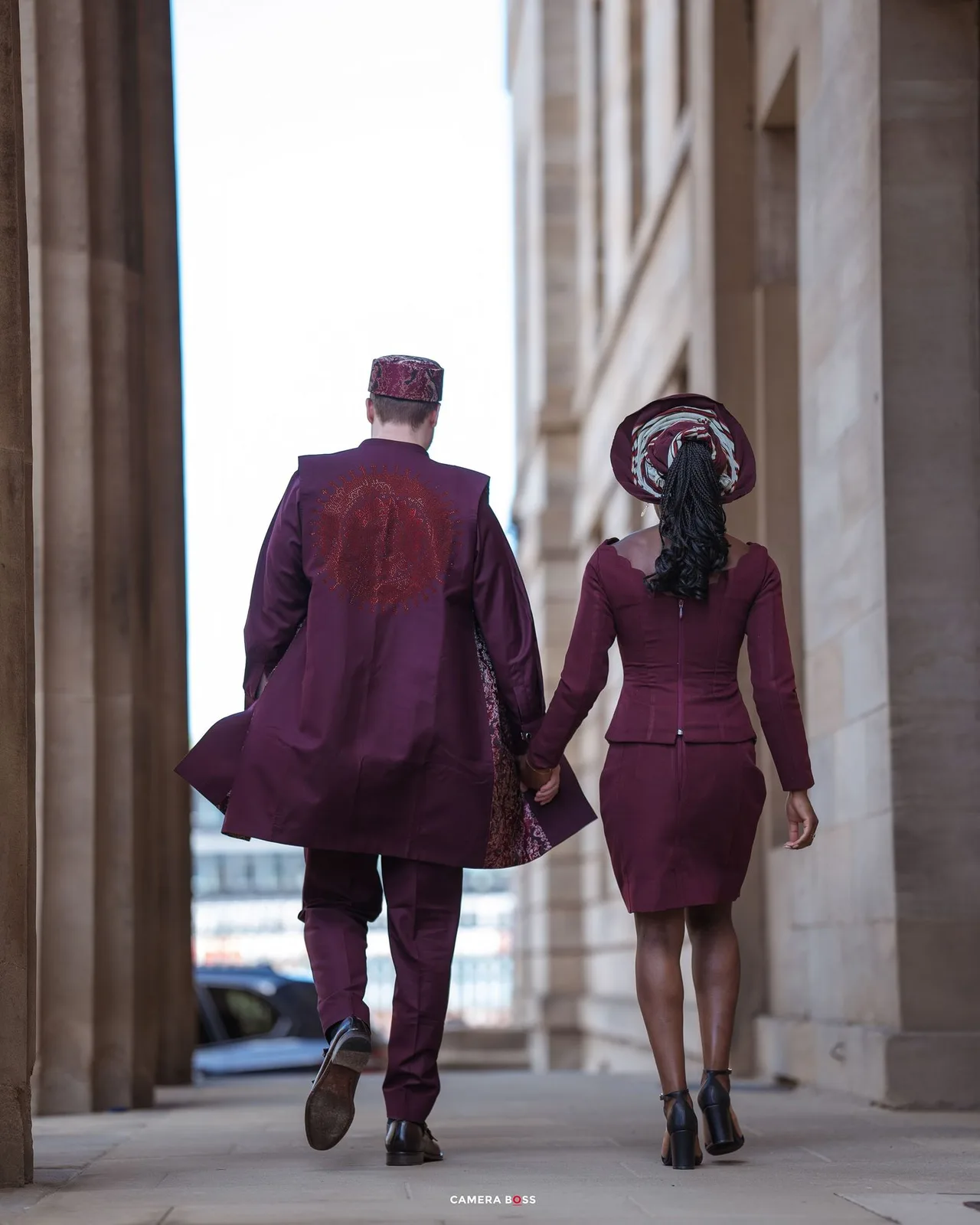 Stylish couple wearing coordinated burgundy outfits walking together between elegant stone columns.