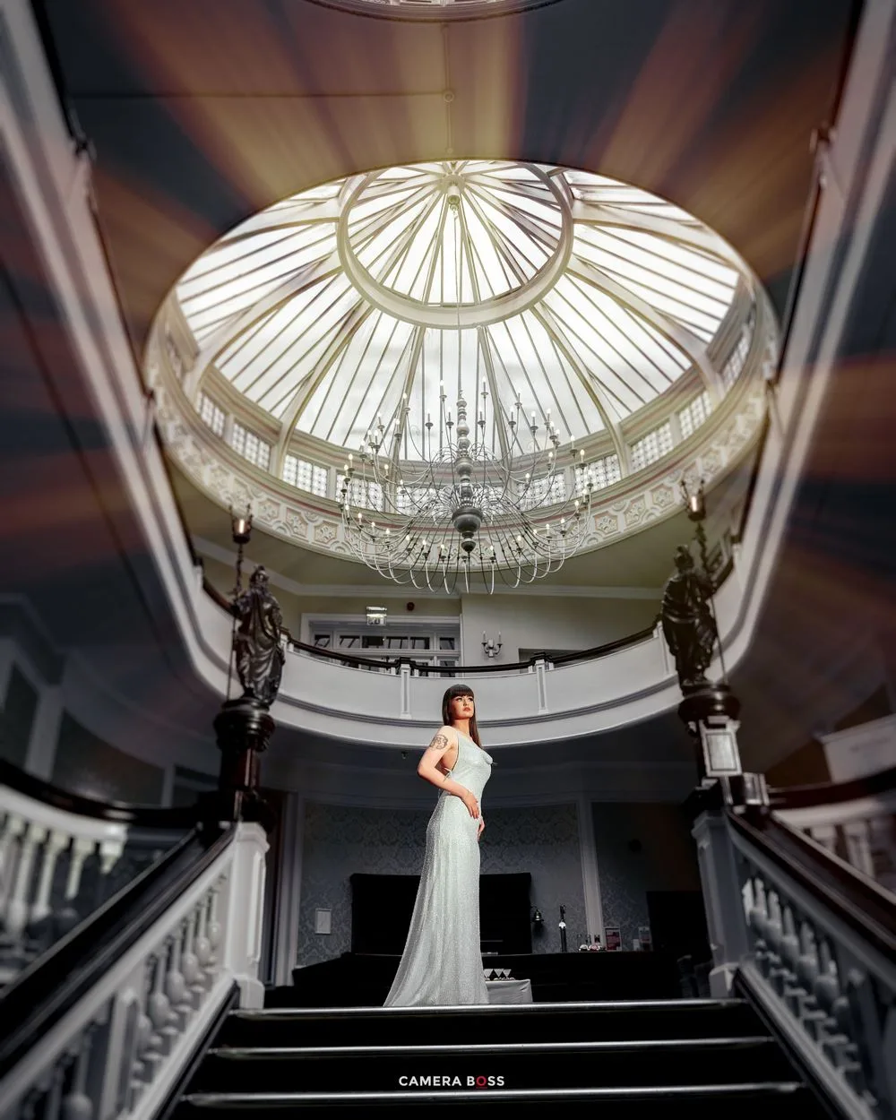 Elegant figure stands on grand staircase under ornate domed glass ceiling with dramatic lighting in historic building.