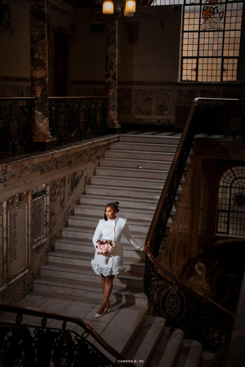 Bride in white short dress holding pink bouquet on grand marble staircase in elegant historic building.
