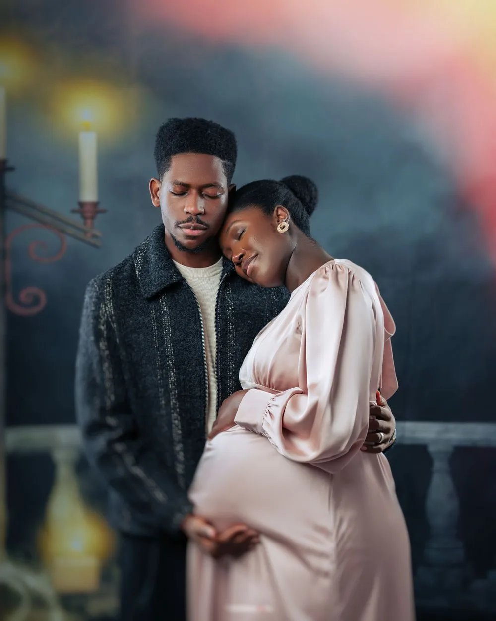 A couple shares an intimate moment during a maternity photoshoot with candlelight and dreamy atmosphere in the background.
