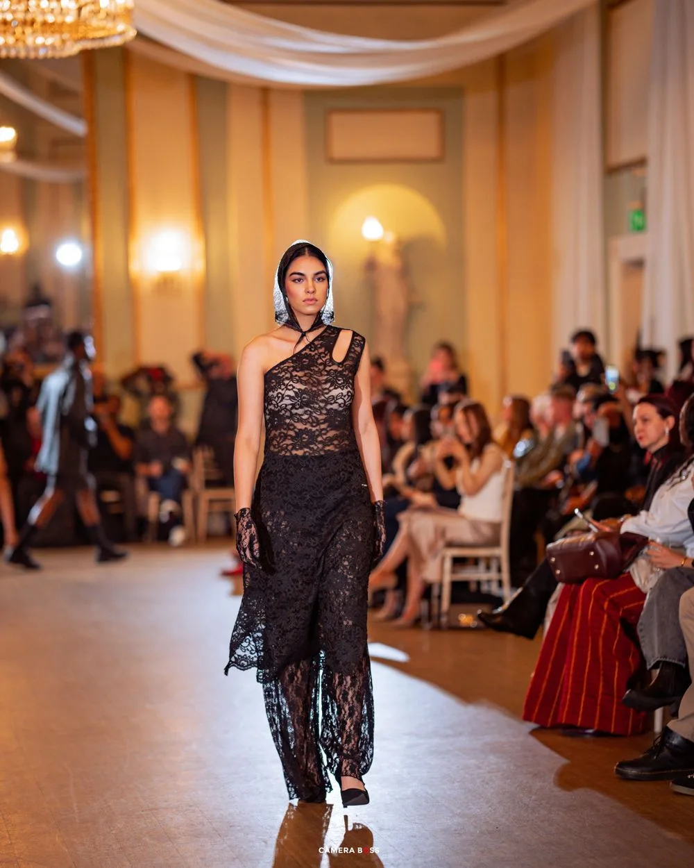 Model wearing elegant black lace one-shoulder evening gown walks runway at fashion show in ornate ballroom setting.