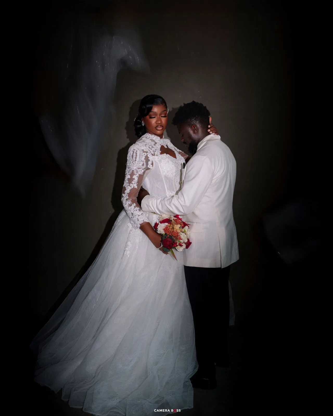 Elegant bride and groom in white wedding attire pose romantically against a dark background holding a bouquet.