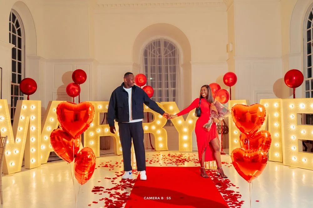 Romantic proposal scene with illuminated LOVE letters, red balloons, heart decorations and rose petals on red carpet.