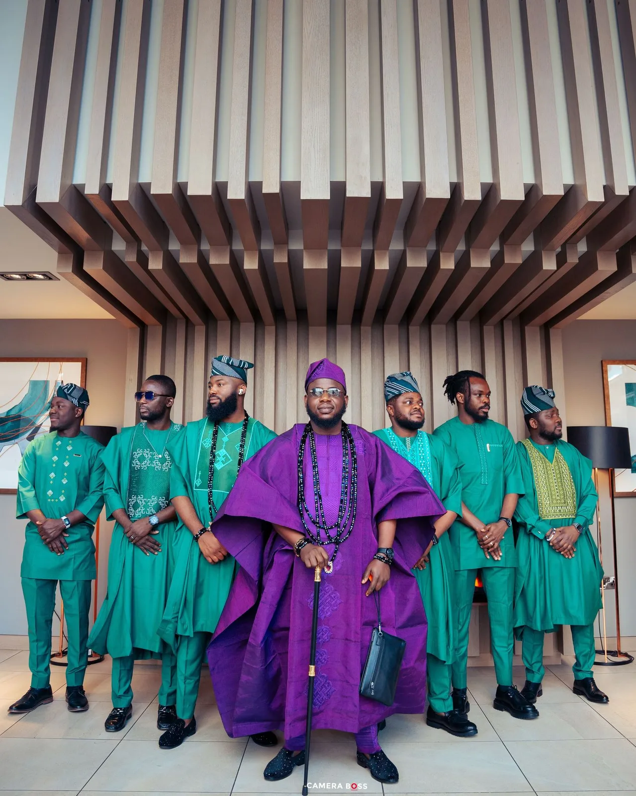 A traditional Nigerian wedding party group wearing coordinated green agbada outfits with one central figure in regal purple attire.