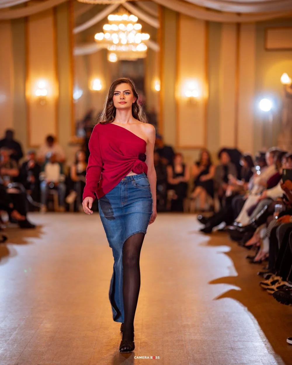 Fashion model walks runway in red one-shoulder top and denim skirt at elegant ballroom fashion show.