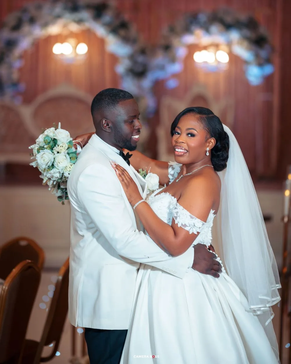 A joyful wedding moment captured in an elegantly lit venue with white decor and warm glowing chandeliers in the background.