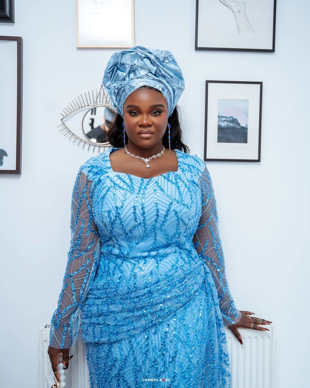Person wearing a vibrant blue lace gown with matching headwrap poses against a white wall with framed artwork.