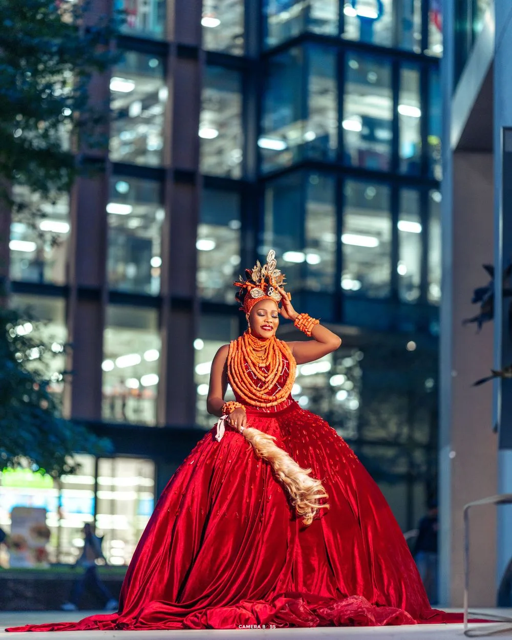 A performer in a stunning red ball gown and ornate headpiece poses dramatically against a modern glass building backdrop.