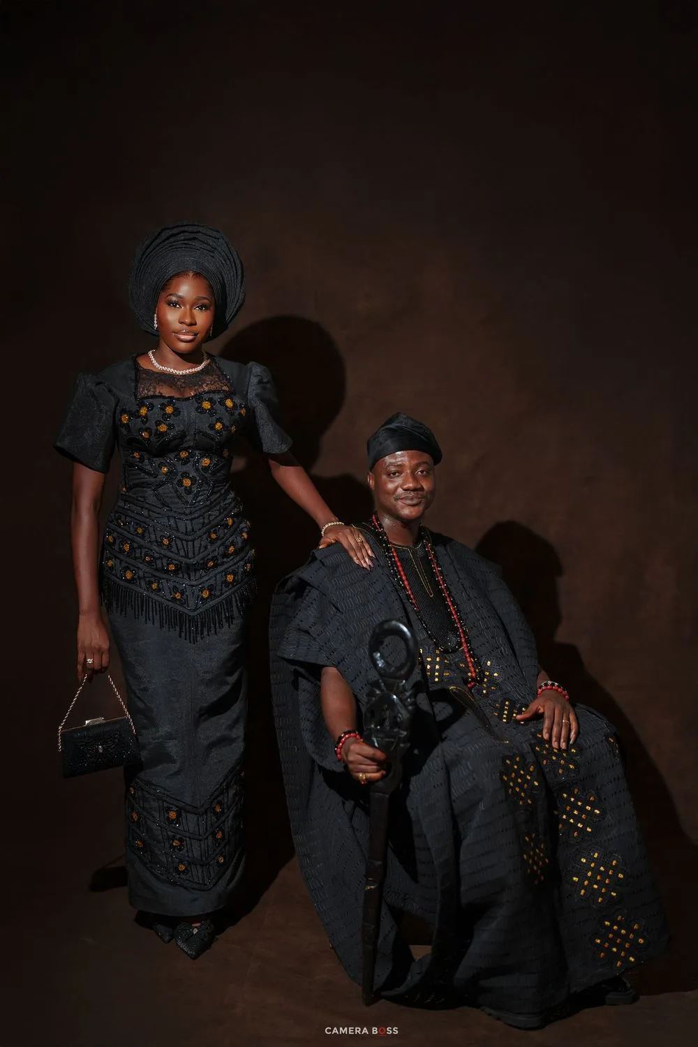Two people in matching dark traditional African attire pose elegantly for a formal portrait photo.