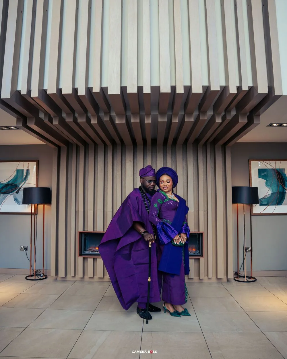 A couple in coordinating purple traditional Nigerian wedding attire poses in front of a modern architectural backdrop.