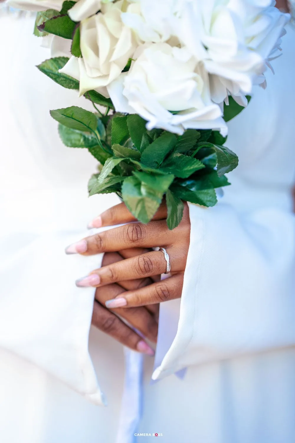 A bride holding white roses in a bridal bouquet showing her wedding ring and manicured nails against white fabric.