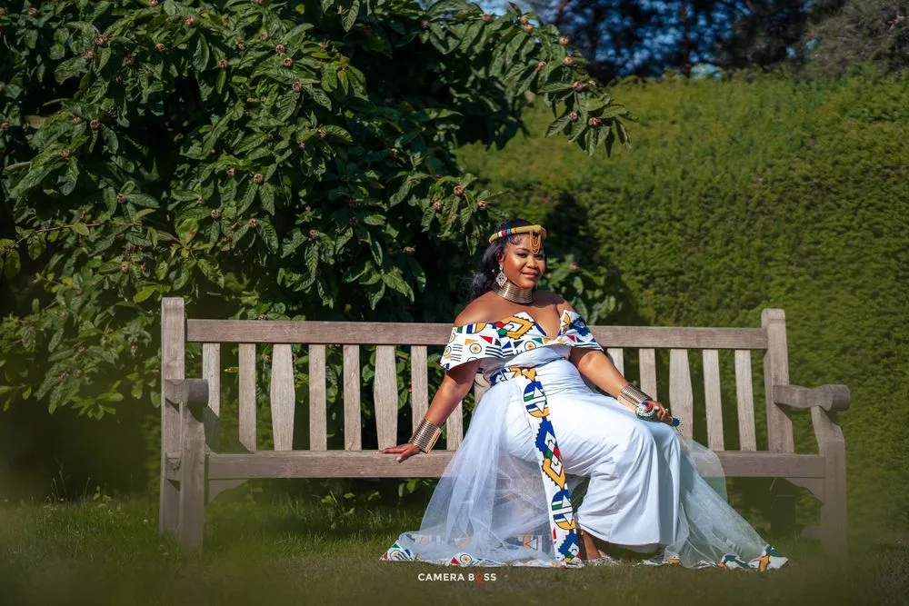 Someone in a flowing light blue gown sits elegantly on a wooden garden bench surrounded by green foliage.