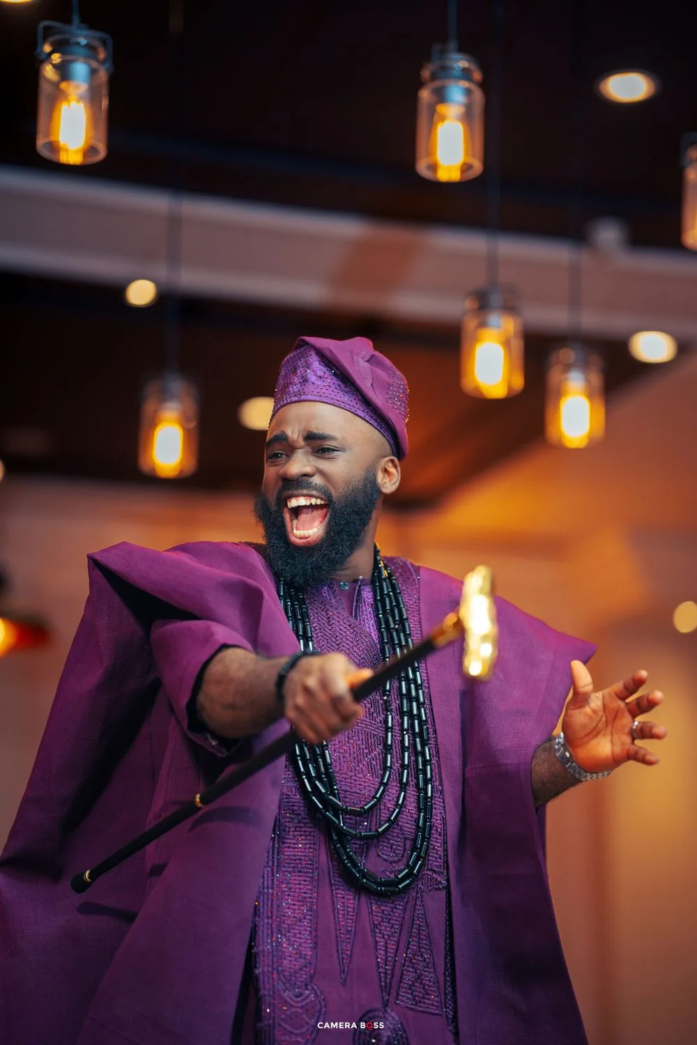 A performer in vibrant purple traditional African attire and beaded necklaces gestures expressively under warm pendant lighting.