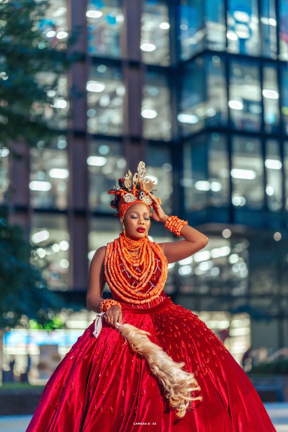 Model poses in traditional Nigerian wedding attire including red ball gown and coral beads against modern office building.