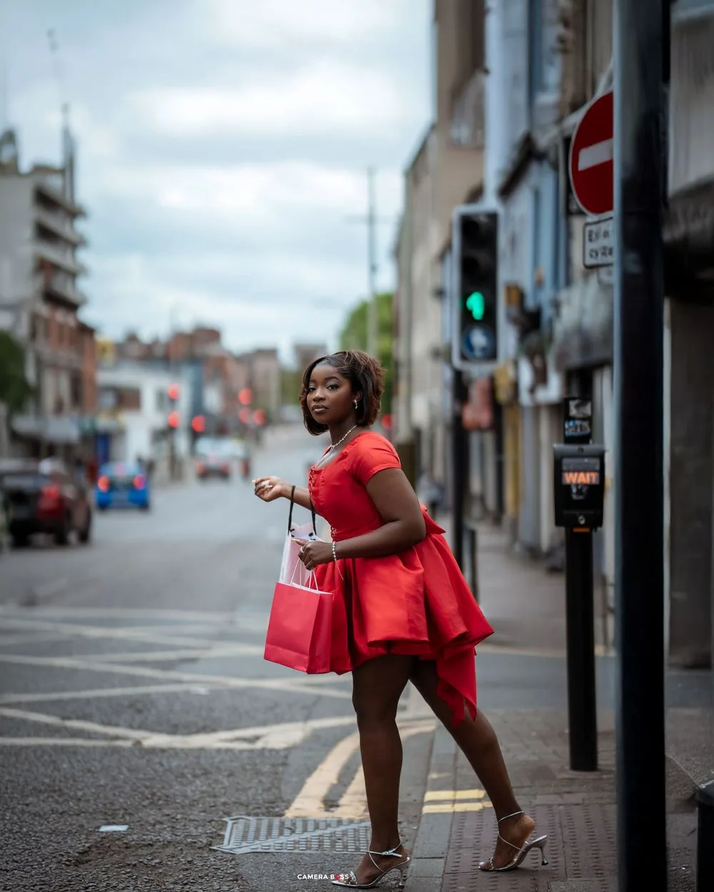 A person in a red outfit carrying shopping bags walks along a quiet urban street with buildings in the background.