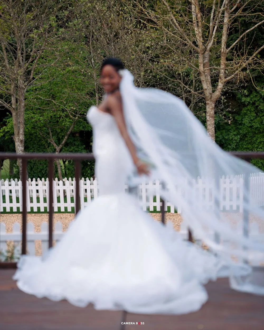 A flowing white wedding gown and veil catch the breeze on a wooden deck surrounded by white picket fencing.