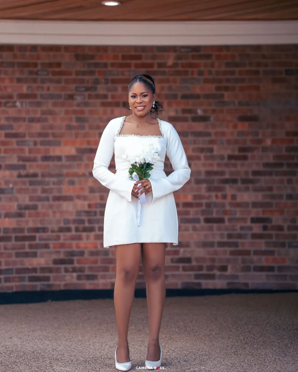 A bride in a short white dress and long sleeves holds a small bouquet while standing against a brick wall.