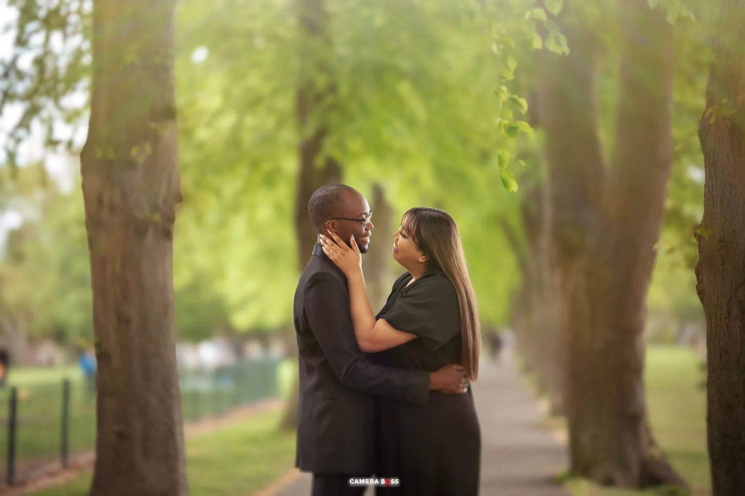 Two silhouettes embrace in a romantic moment along a tree-lined path with soft bokeh lighting in the background.