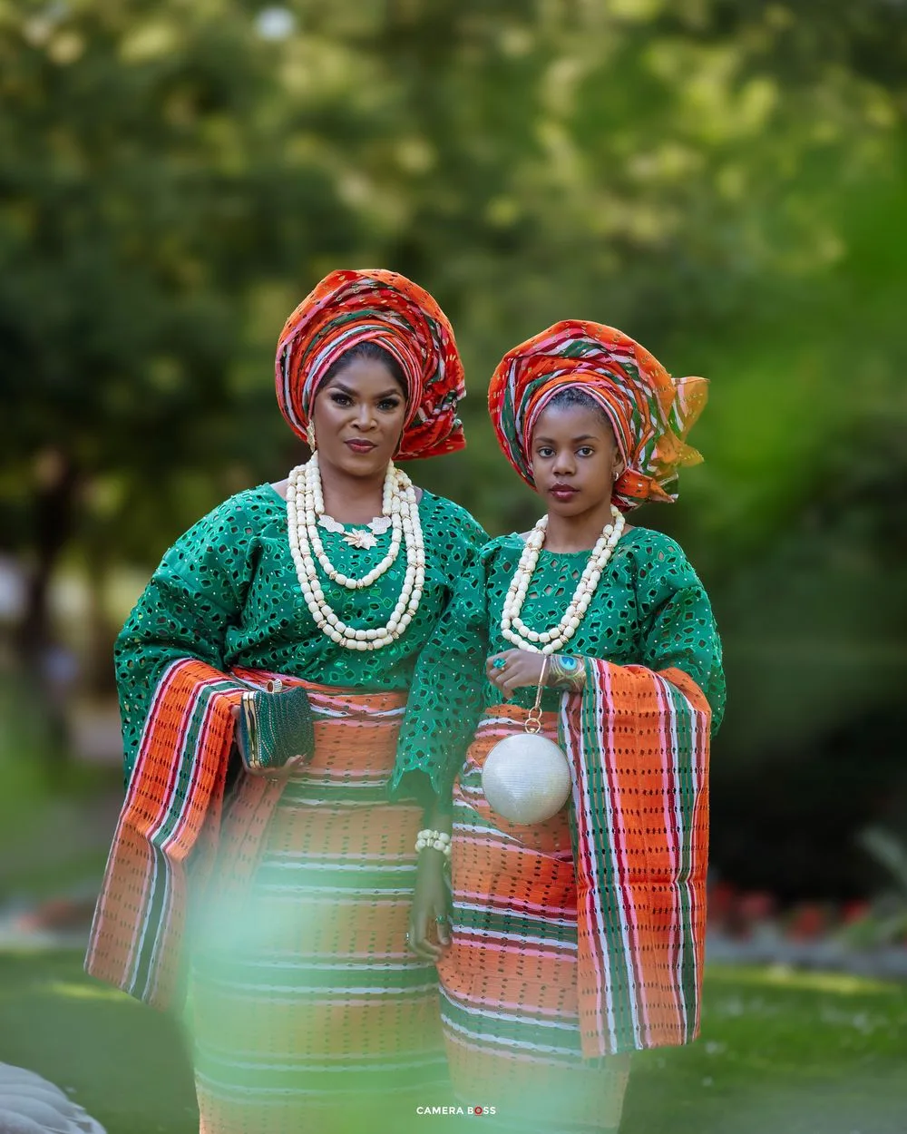 Two people wearing traditional African attire with colorful headwraps, green lace tops and striped wrappers pose in a garden setting.