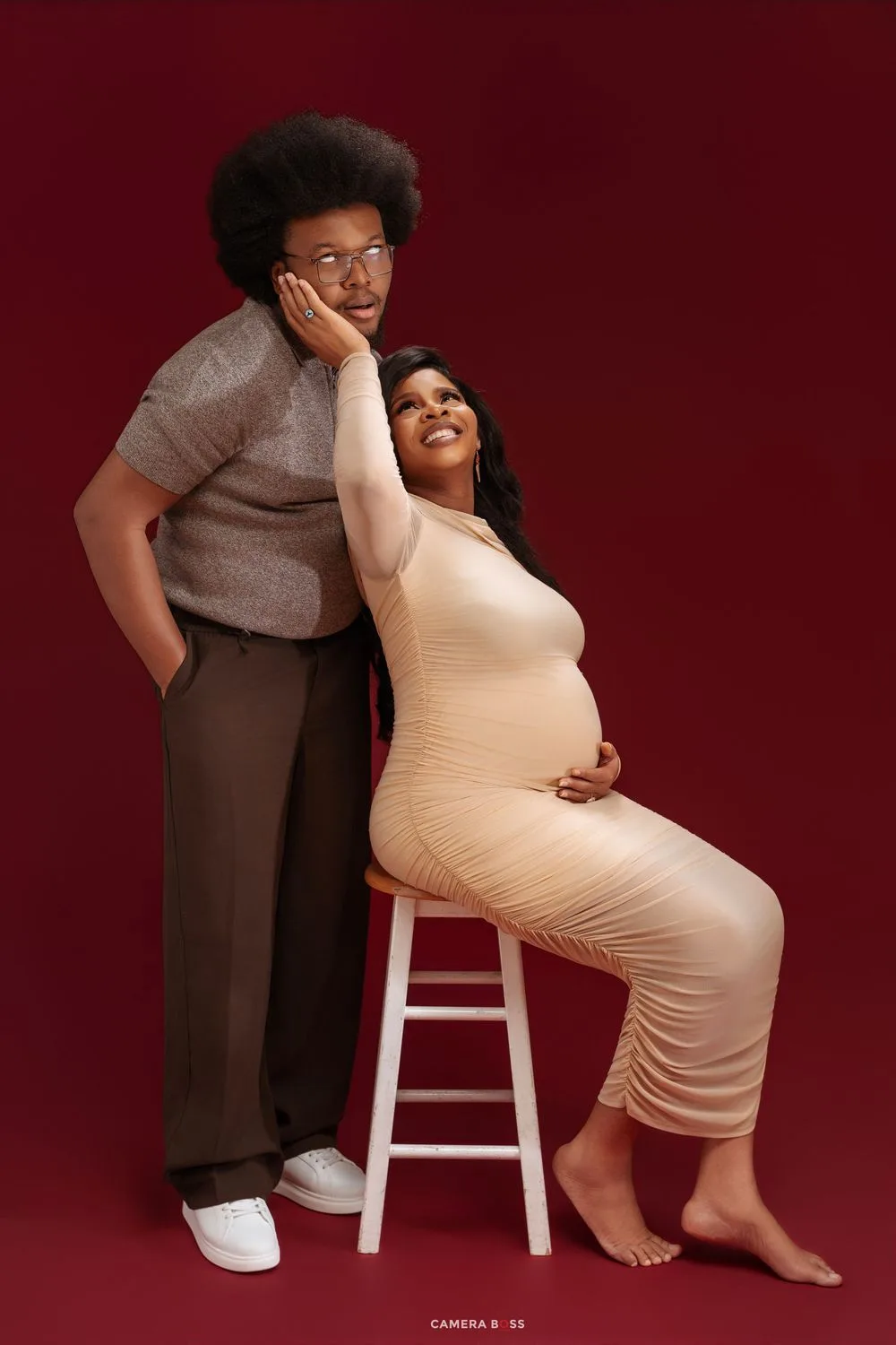 Couple maternity photo with pregnant woman in nude dress seated on stool leaning on partner against red backdrop.