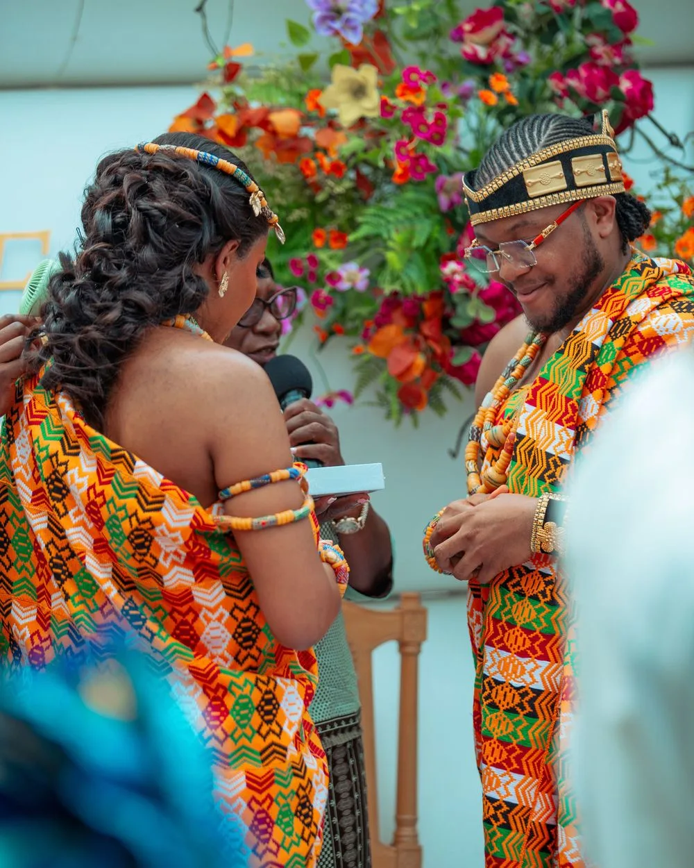 Two people in vibrant traditional African kente cloth attire exchange vows during a wedding ceremony with colorful flowers in background.