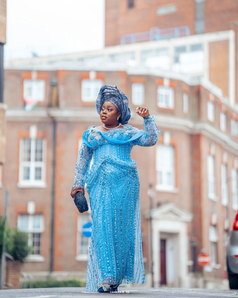 Someone in a sparkling blue formal dress and matching headwrap poses elegantly outside a brick building.