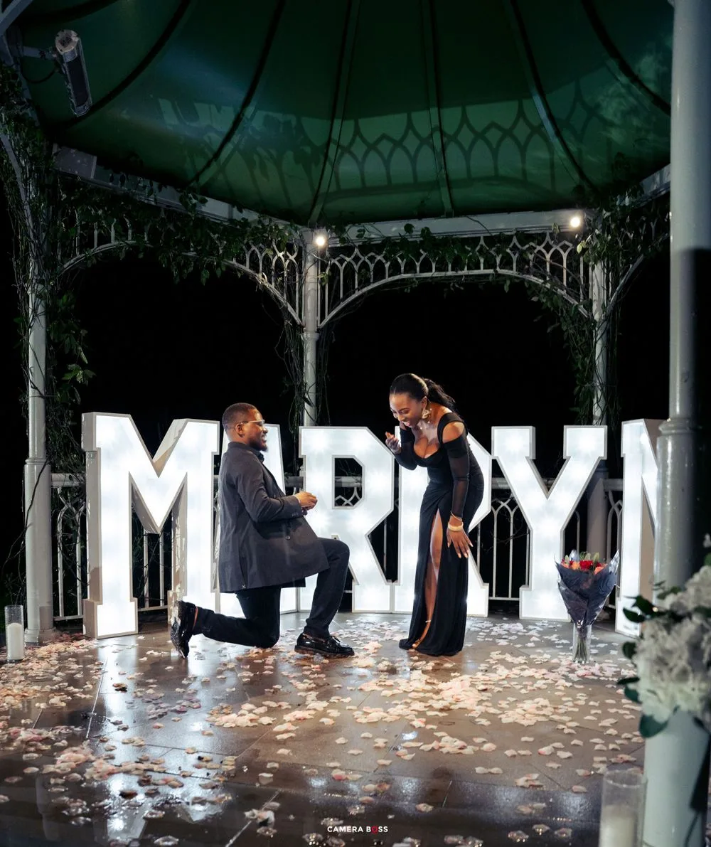 A romantic marriage proposal scene under a lit gazebo with large illuminated 'MARRY' letters and scattered rose petals.