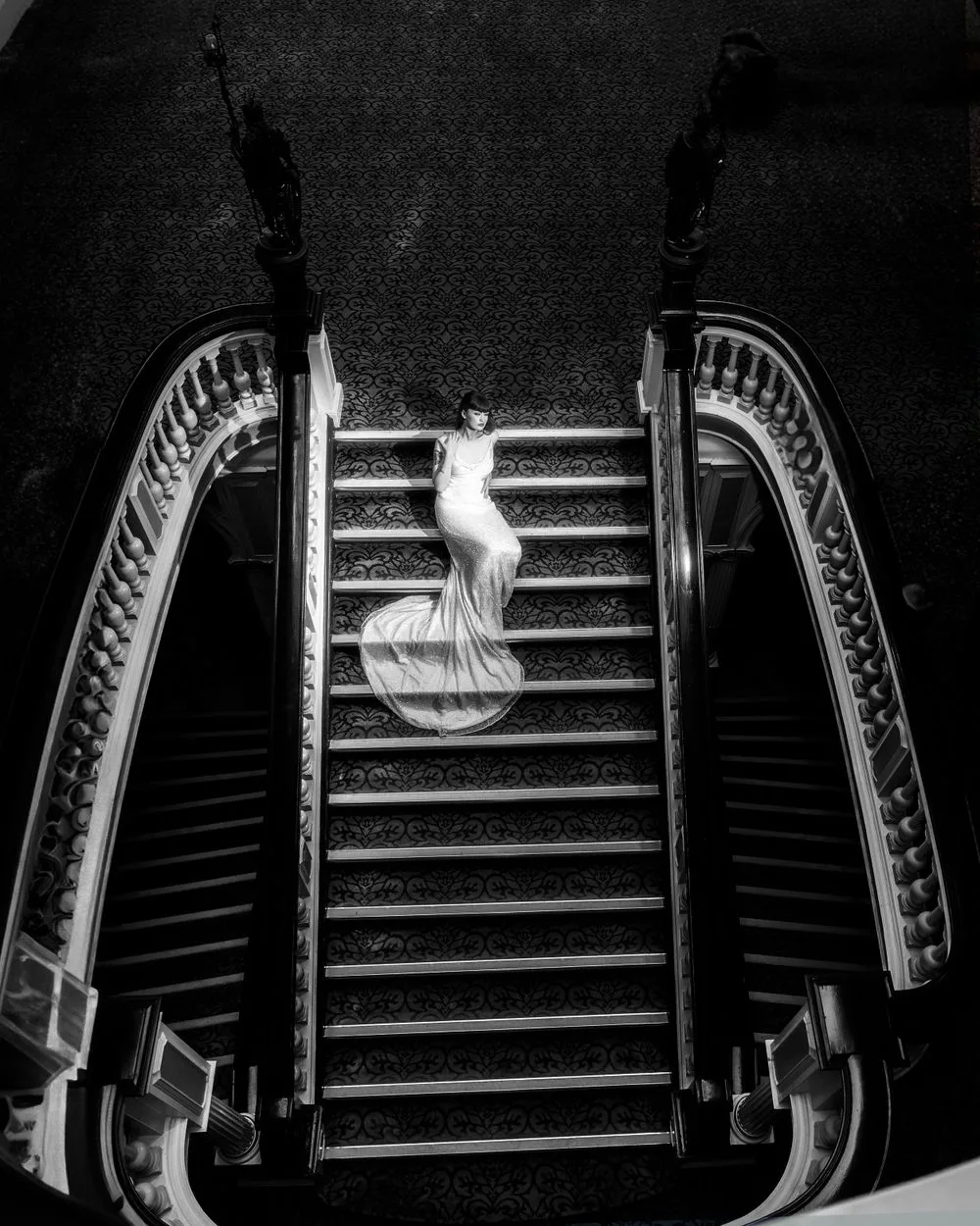 A dramatic black and white photograph of a figure in a flowing dress ascending an elegant curved staircase.