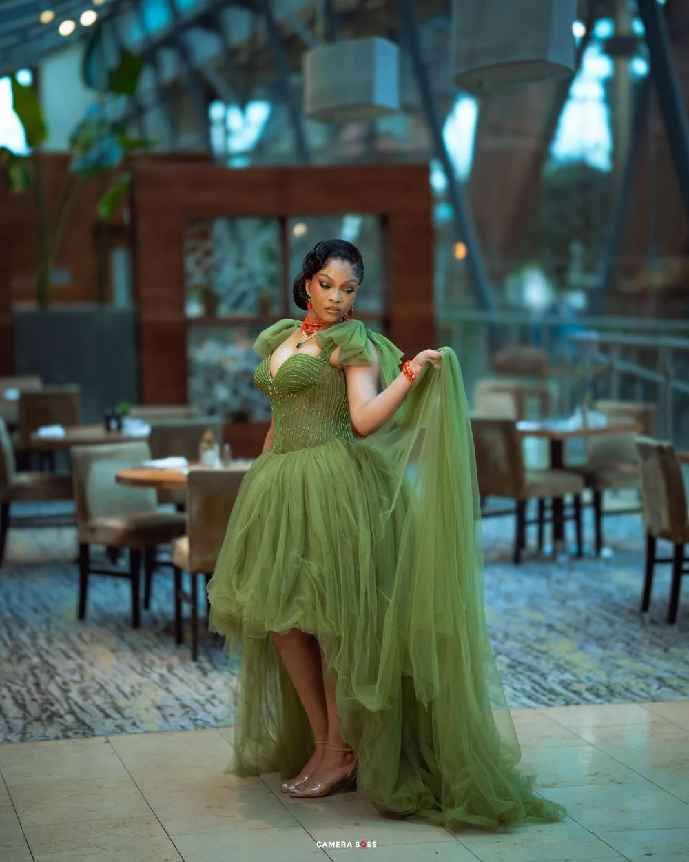 A woman poses gracefully in a flowing olive green tulle gown at an elegant restaurant setting with soft lighting.