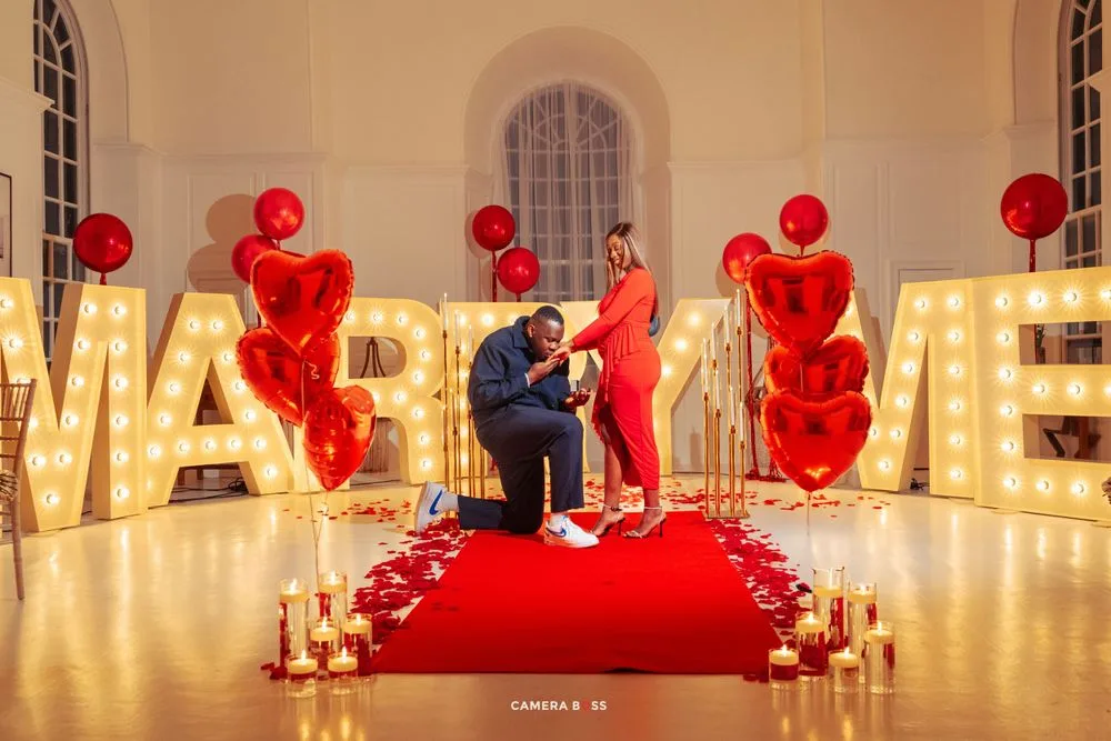 Illuminated marquee 'MARRY ME' lettering with red balloons and candles on red carpet for romantic proposal setup.