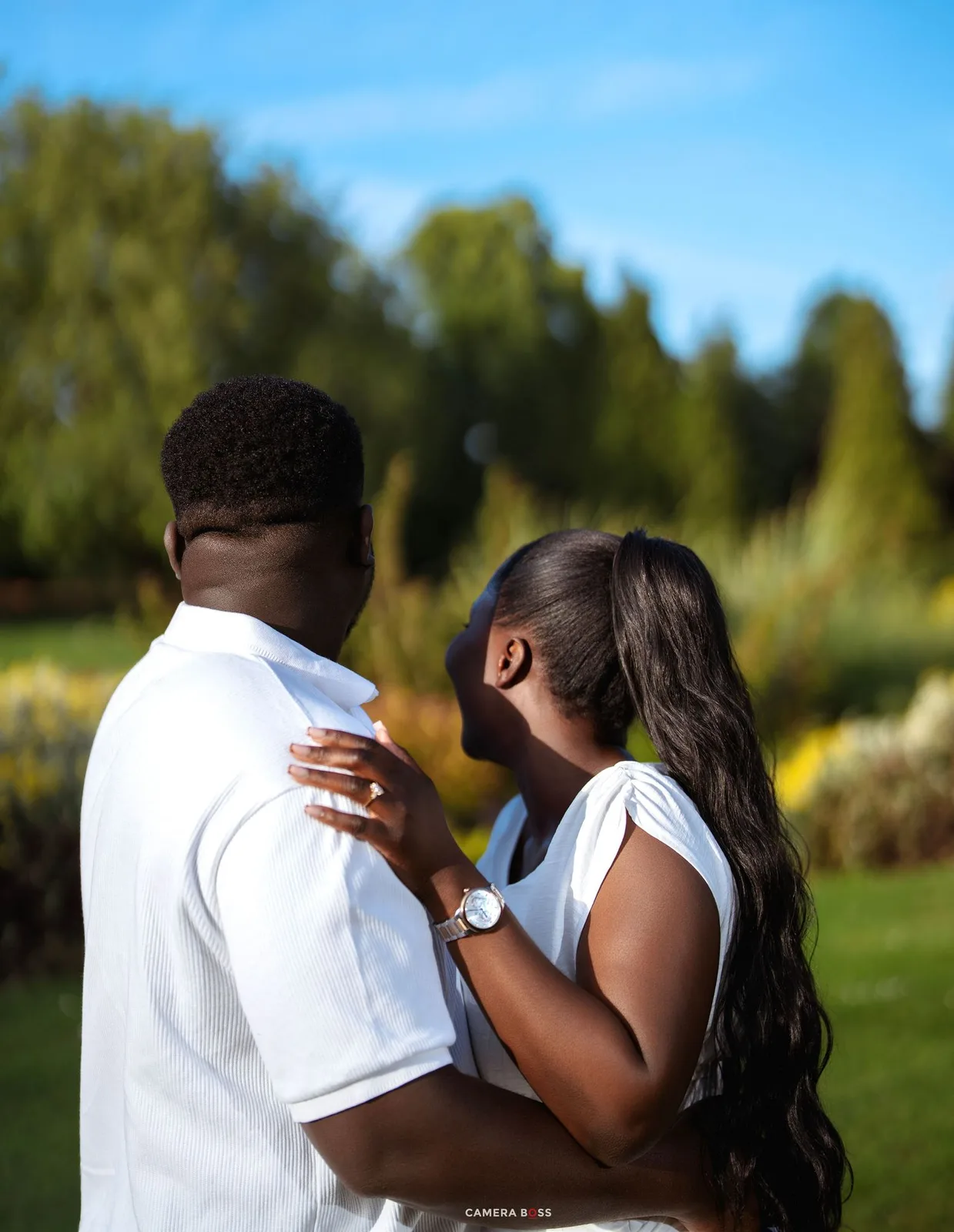 A couple in white outfits embraces outdoors in a lush green garden on a sunny day.